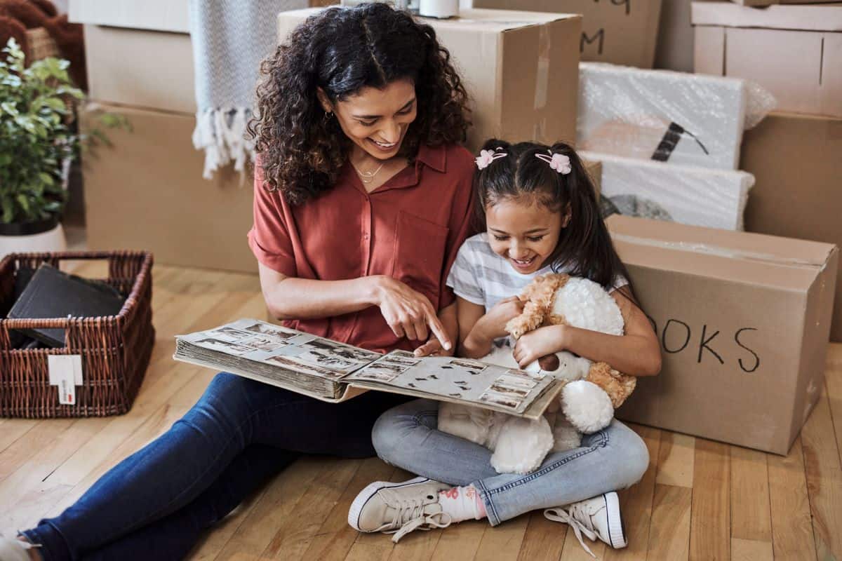 Cropped mother and daughter sitting on the floor looking at a photo album among moving boxes.